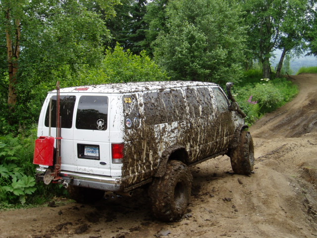 White van covered in mud, driving on a dirt trail with trees in the background. Has a red gas can and shovel mounted on the back
