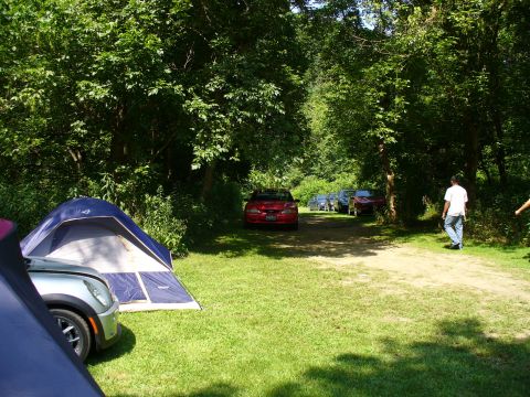 1991 Ford Probe - A red car is parked on a dirt road surrounded by trees and tents, with other vehicles visible in the background.