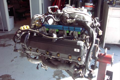 1992 Ford Crown Victoria - Modular overhead-cam Ford V8 sitting on a red engine stand in a shop, fitted with a blue anodized fuel rail, green-tipped injectors, cast i…