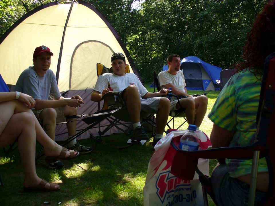 People relax in camping chairs near tents, with a large bag and water bottle in the foreground