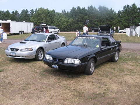 1989 Ford Mustang - Two Ford Mustangs are parked on a grassy area with trees and trailers in the background.