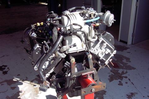 1992 Ford Crown Victoria - A V8 engine sitting on a red engine stand outside a shop bay, fitted with a supercharger and intake plenum on top, a remote reservoir on a…