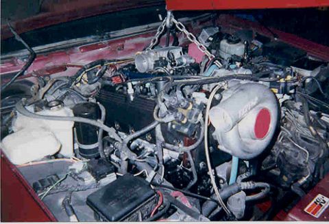 1992 Ford Crown Victoria - An engine hangs from a hoist chain as it is lowered into the bay of a red car, with a white round air cleaner housing, black valve cover, d…