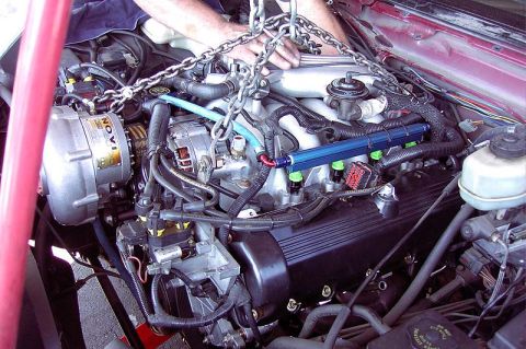 1992 Ford Crown Victoria - A supercharged V8 is lowered into a red car's engine bay on a chain hoist, showing a belt-driven blower, an anodized blue fuel rail and exp…