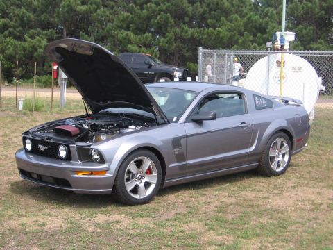 1989 Ford Mustang - A gray Ford Mustang is parked on grass with its hood open, revealing the engine compartment. A black truck is visible in the background.