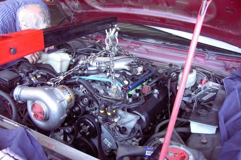 1992 Ford Crown Victoria - An engine is being lowered into the bay of a maroon car on chains and a load leveler, with a person in a blue striped shirt steadying the r…