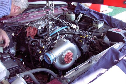 1992 Ford Crown Victoria - A Ford V8 engine hangs from a hoist chain being lowered into the engine bay of a maroon car, with a large polished Vortech centrifugal supe…