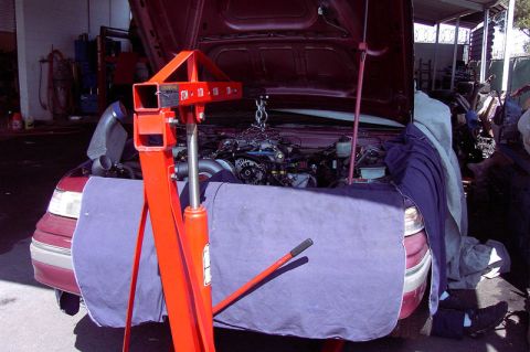 1992 Ford Crown Victoria - An orange 2-ton hydraulic engine hoist positioned over the open engine bay of a dark red sedan, chain hooked to an engine being lowered int…