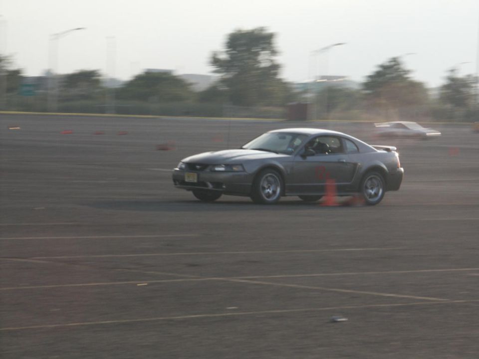 Gray Ford Mustang on a track, driver visible, orange cones nearby. Another car is in the background