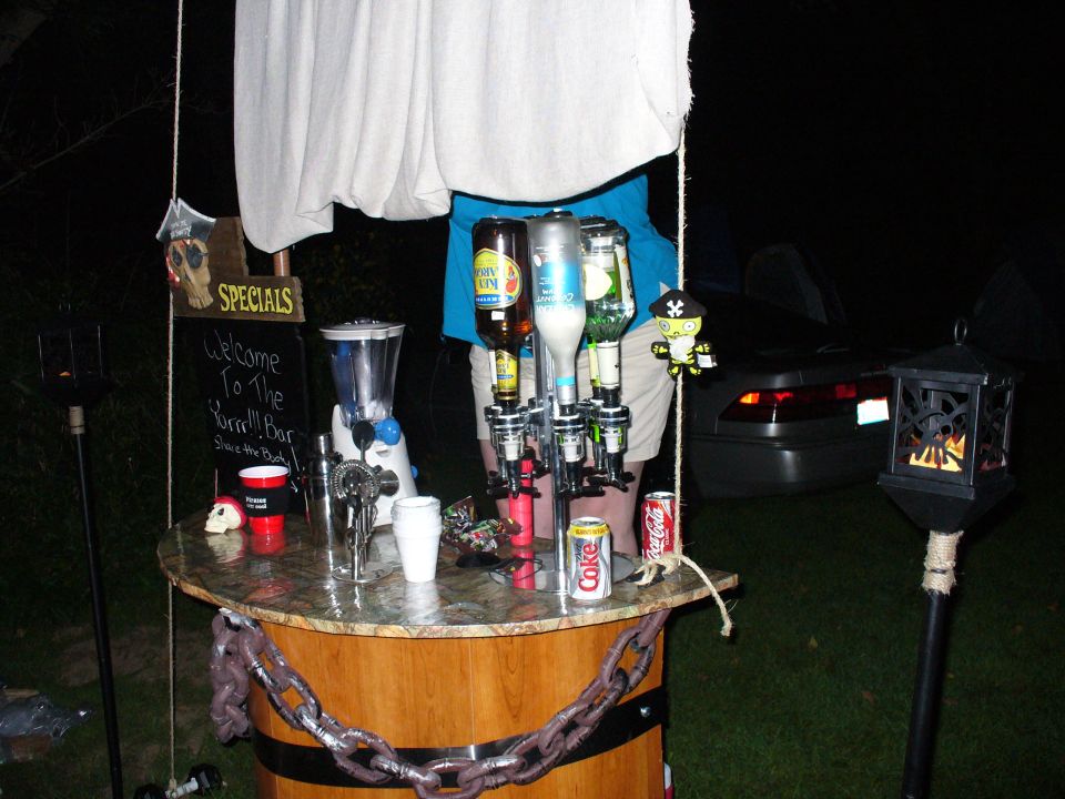 Outdoor bar with bottles, blender, and cups. A car is parked behind it at night