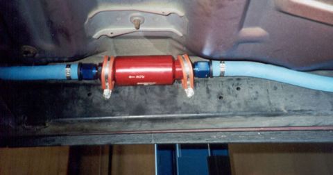 1992 Ford Crown Victoria - A red anodized inline fuel filter marked with a 'FLOW' directional arrow, mounted along a frame rail and plumbed with blue braided fuel lin…