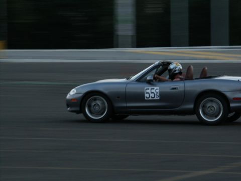 2001 Mercury Grand Marquis - A gray Mazda Miata convertible with racing number 55 speeds around a track, driver in helmet.