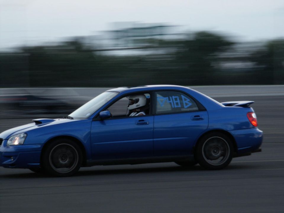 Blue Subaru Impreza WRX sedan with racing numbers on the window, driving on a track. Another car is blurred in the background
