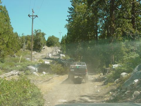 1986 Ford Bronco - A pickup truck kicks up dust on a rough dirt road surrounded by trees and rocks.
