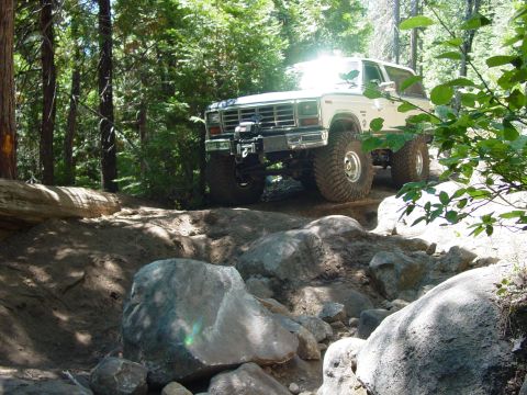1986 Ford Bronco - This lifted SUV navigates a challenging, rocky off-road path through a wooded area.