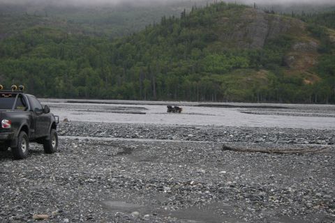 2003 Jeep TJ - A black pickup truck is parked on a gravelly riverbed, with another vehicle partially submerged in the water in the distance.