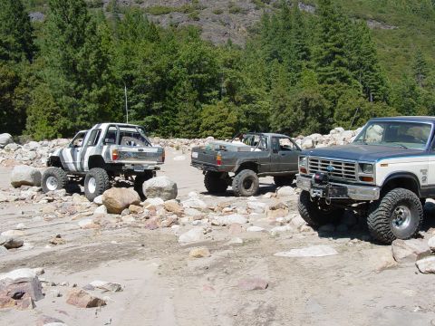 1986 Ford Bronco - Three lifted 4x4s staged on a rocky, sandy flat below a granite ridge and pines: a white Toyota mini-truck with a chopped cab, a gray Toyot…