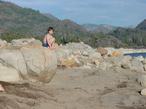 1986 Ford Bronco - A young boy sits on rocks overlooking a lake with mountains in the distance. The image has a watermark for Supermotors.net.