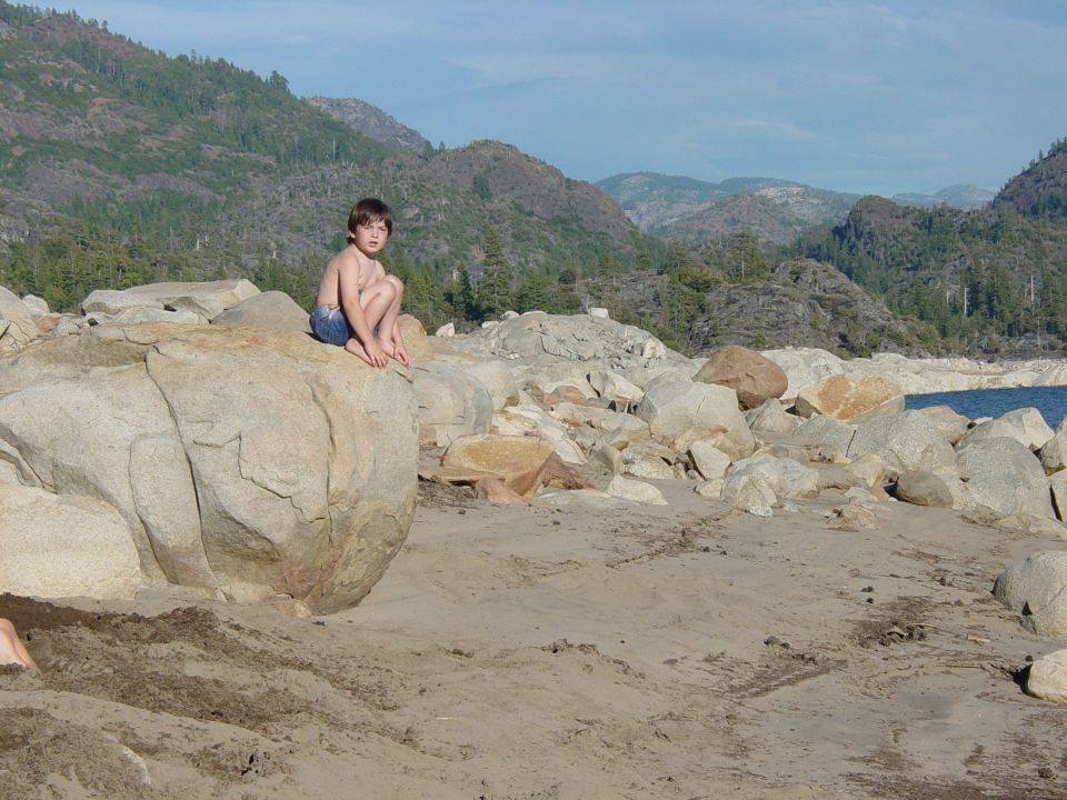 Boy sits on large rocks near a body of water with mountains in the background. Watermark reads www.supermotors.net