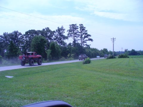 1996 Ford Bronco - A red lifted truck drives down a rural road, followed by other vehicles under a cloudy sky.