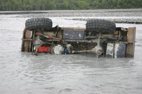 2003 Jeep TJ - An overturned vehicle rests submerged in a river, its undercarriage and suspension system exposed to the murky water.