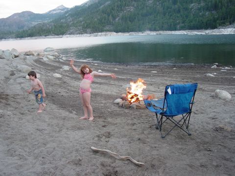1986 Ford Bronco - Two kids in swimsuits play on the sandy shore of a mountain lake beside a rock-ringed campfire and a blue folding camp chair, with tire tra…