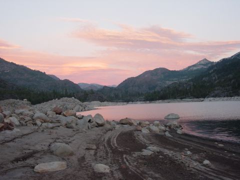 1986 Ford Bronco - Pink sunset over a mountain lake ringed by granite boulders and conifer-covered ridges, with fresh tire tracks cut into the wet sand along…