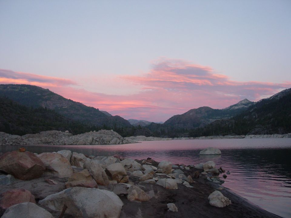 Pink sunset over a granite mountain lake with boulder shoreline and forested ridges