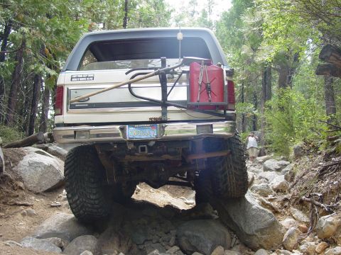 1986 Ford Bronco - Rear view of a white lifted Ford SUV straddling boulders on a forest rock trail, with the rear axle, leaf springs and driveshaft fully expo…