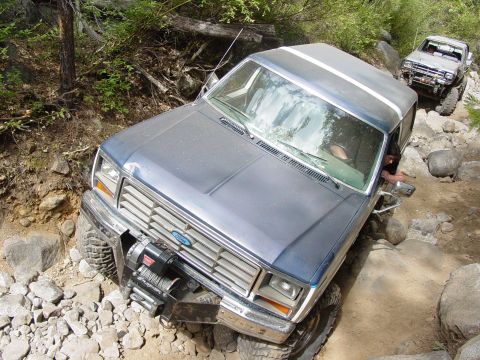1986 Ford Bronco - Elevated front three-quarter view of a dusty blue Ford 4x4 with a white roof working down a rocky trail, body leaning as the front wheels d…