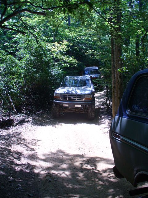 1996 Ford Bronco - Two Ford Broncos navigate a wooded trail on a sunny day, with a third vehicle partially visible.