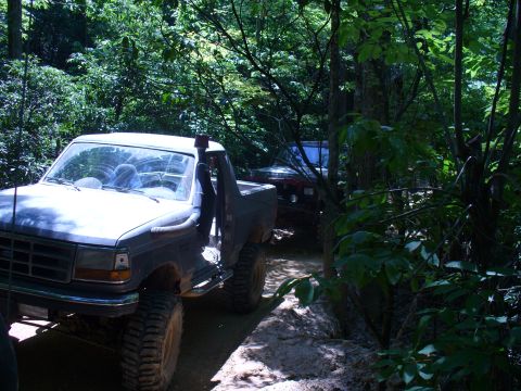 1996 Ford Bronco - Two off-road vehicles navigate a wooded trail, with the lead Bronco showing modifications for rough terrain.