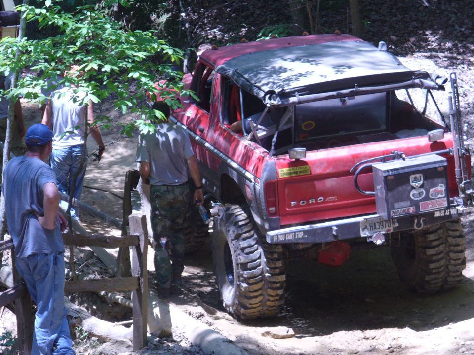 Red Ford Bronco with lifted suspension and large tires on a dirt trail, people standing nearby
