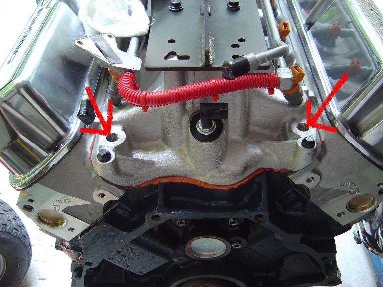 V8 engine valley with intake off, chrome valve covers, red arrows on two bolt holes