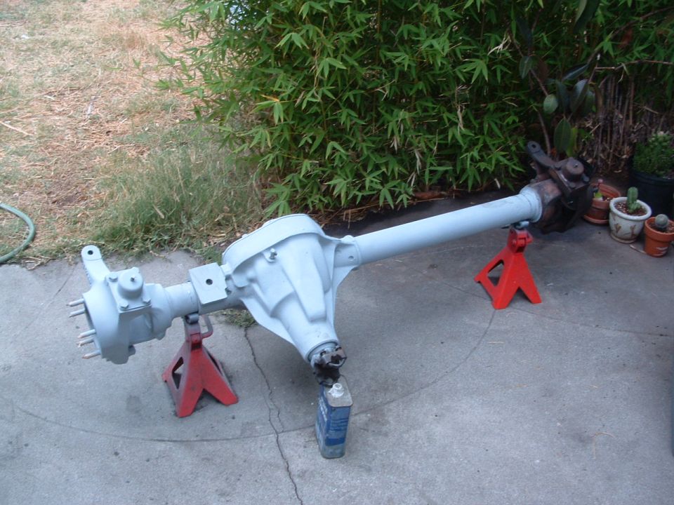 Rear axle assembly on red jack stands, with a bottle of fluid nearby. Bamboo and potted plants in background