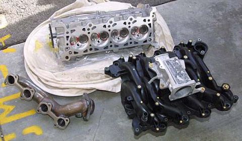 1992 Ford Crown Victoria - Modular V8 engine parts laid out on a concrete floor with yellow parking-stripe paint: an aluminum SOHC cylinder head resting on a white cl…