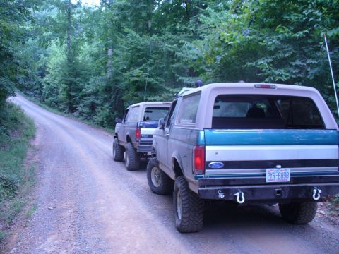 1996 Ford Bronco - Two Ford Broncos drive down a wooded dirt road, with the lead vehicle partially obscured.