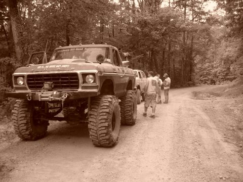 1996 Ford Bronco - A heavily modified Ford F-Series truck with oversized tires and a winch sits on a dirt road, with several people standing nearby.