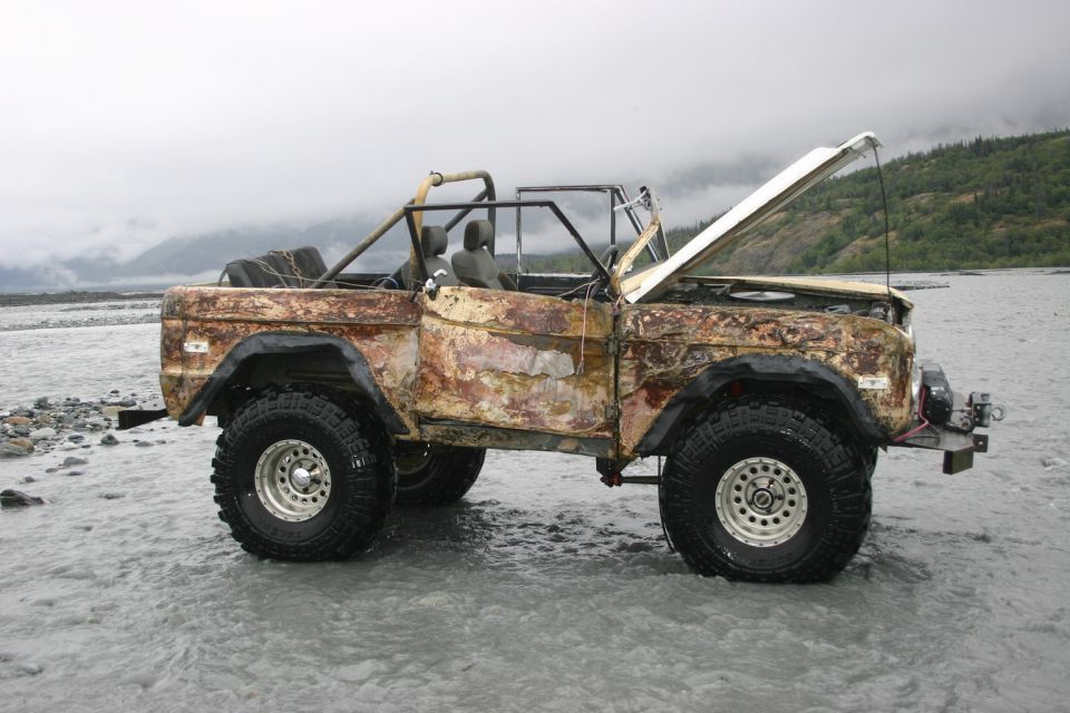 Rusty 1970s Ford Bronco with hood open, parked in shallow water