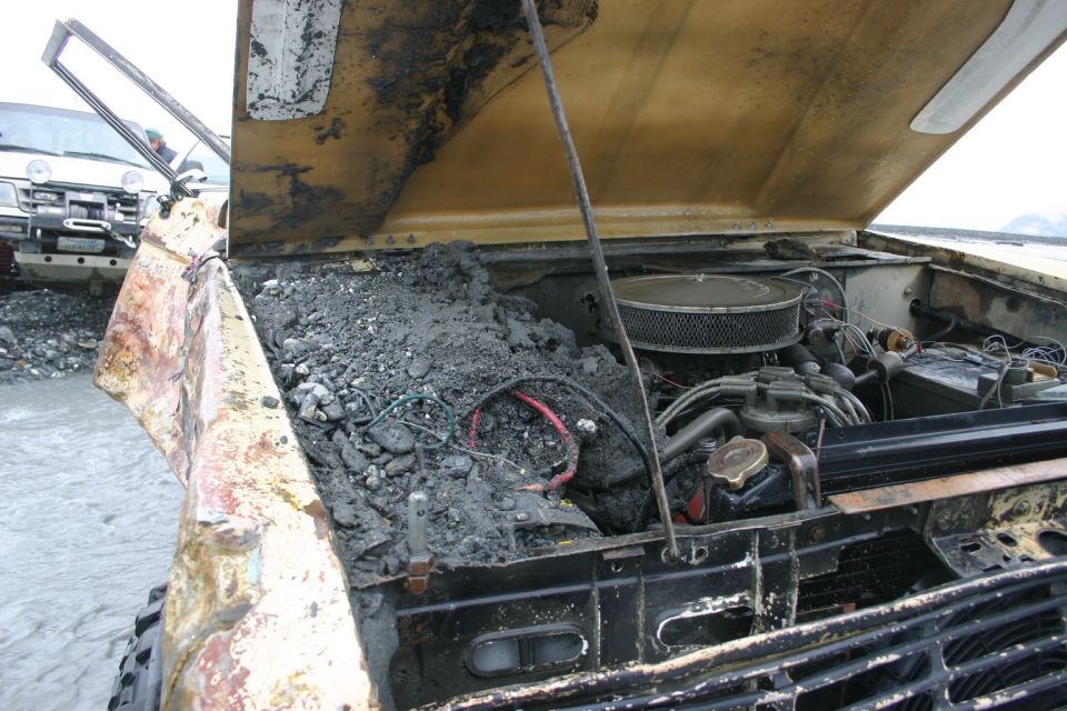 Engine bay of a vehicle is full of mud and debris. Another vehicle is visible in the background
