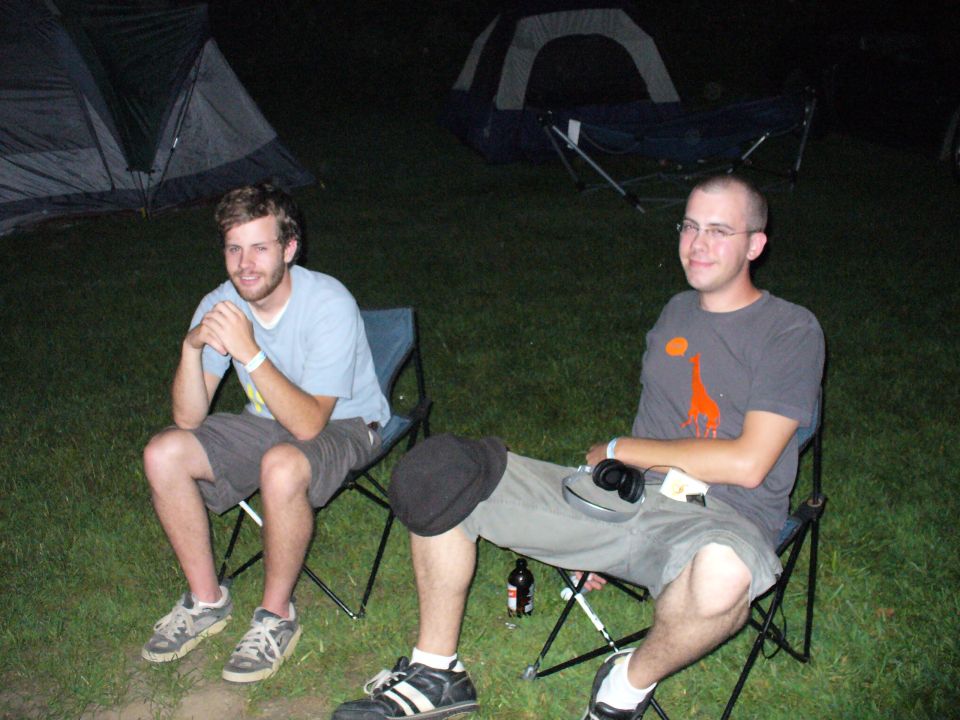 Two men sit in camping chairs outdoors at night with tents in the background
