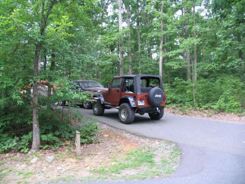 1995 Ford C-350 (Centurion Conversions) - A brown Jeep Wrangler is parked on a paved road in a wooded area, with a maroon SUV behind it.