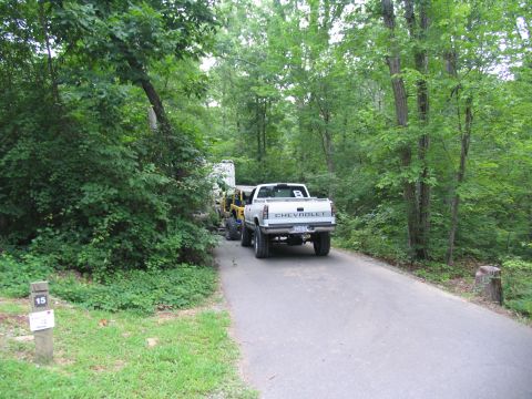 1995 Ford C-350 (Centurion Conversions) - Chevrolet pickup truck towing a trailer with a Jeep on a paved road in a wooded area. Site 15 marker visible.