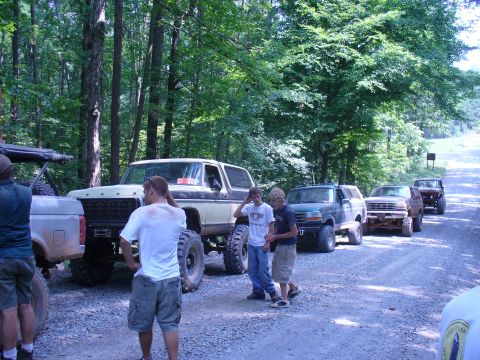 1996 Ford Bronco - A group of off-road enthusiasts and their modified vehicles are gathered on a dirt road surrounded by trees.