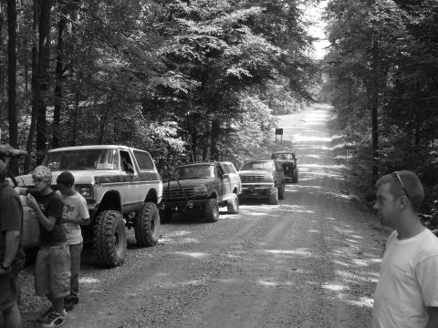 1996 Ford Bronco - A group of off-road vehicles, including a lifted 1996 Ford Bronco, travel down a wooded dirt road.