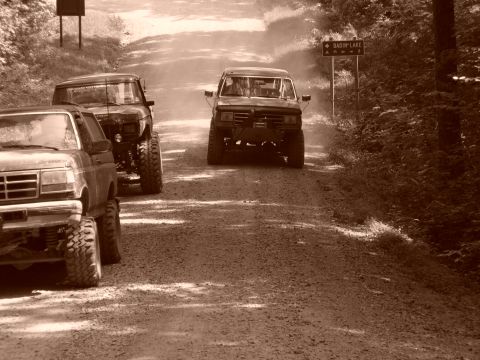 1996 Ford Bronco - Three lifted trucks navigate a dusty dirt road in a wooded area.