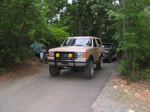 1995 Ford C-350 (Centurion Conversions) - A tan Ford Bronco with custom smiley face lights on its grille guard is parked on a paved road, with another vehicle behind it.