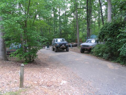 1995 Ford C-350 (Centurion Conversions) - Two trucks are parked at a campsite in a wooded area, with camping gear set up.