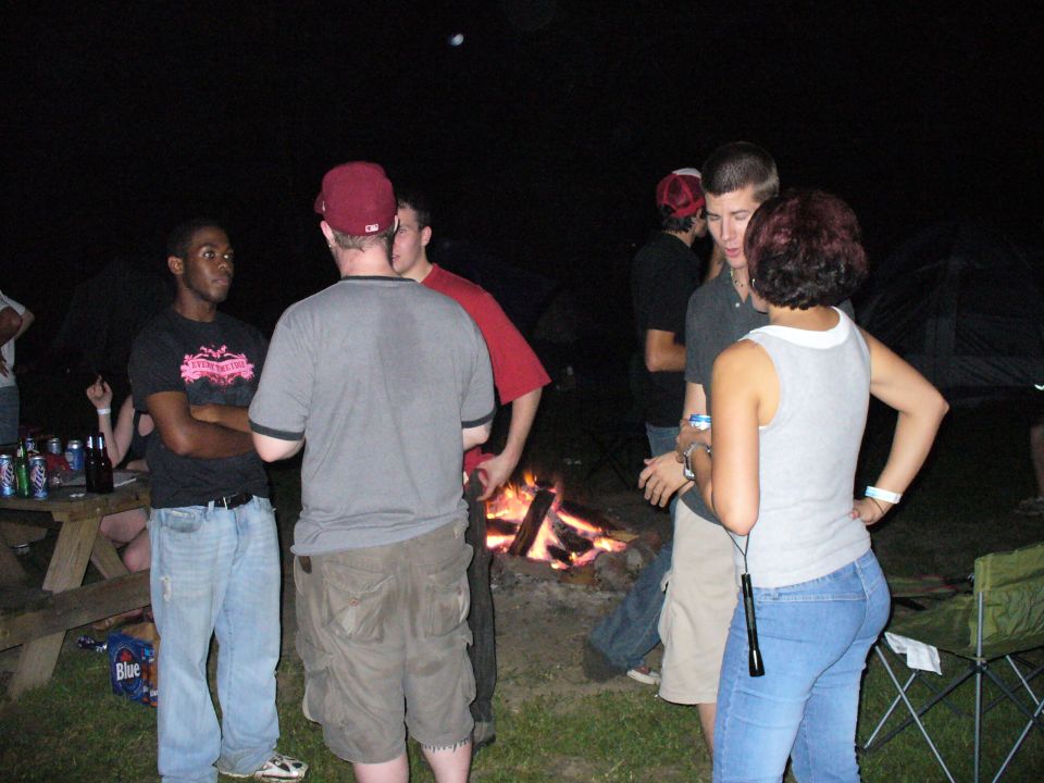 group of people standing around a campfire at night, some holding drinks