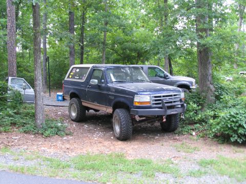 1995 Ford C-350 (Centurion Conversions) - A lifted blue Ford Bronco is parked in a wooded area with another truck nearby.
